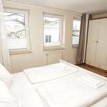 Schlafzimmer mit Doppelbett, Kleiderschrank und Fenstern mit Blick auf Gebäude.