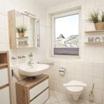 Badezimmer mit Waschbecken, Toilette und Fenster. Holzmöbel und Pflanze auf Regal.