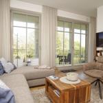 Wohnzimmer mit Sofa, Sessel, Couchtisch und TV. Große Fenster mit Blick auf Balkon.