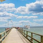 Holzbrücke führt über das Meer, Boot fährt vorbei, Menschen spazieren entlang.