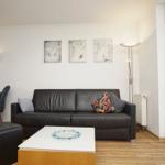 Wohnzimmer mit Sofa, Couchtisch und großen Fenstern. Wandbilder und Stehlampe sichtbar.