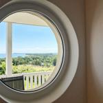 Rundes Fenster zeigt Blick auf Balkon, Grünflächen und Meer.