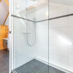 Dusche mit Glaswand, weiße Fliesen und Holzdeckenkonstruktion. Toilettenbereich mit Holzumrandung.