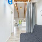 Ein Raum mit grauem Sofa, grauen Vorhängen und einer blauen Lampe. Holzbalken und ein Wandbild sind sichtbar.