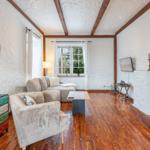 Living room with wooden floor, sofa, armchair, and wall clock. Window with garden view.