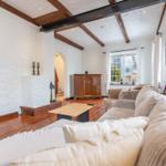 Living room with wooden floor, sofa, TV, and fireplace. Beams and window visible.