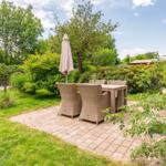 Terrace with table, chairs, and closed umbrella. Greenery and garden house in background.