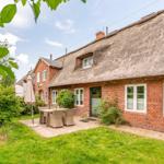 House with thatched roof, garden with table and chairs, green grass and trees.