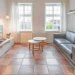 Ein Wohnzimmer mit Sofa, Fernseher und zwei Fenstern. Der Boden ist mit Fliesen ausgelegt.