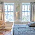 Schlafzimmer mit Bett, Fenstern und Stuhl. Fenster zeigen Außenansicht.