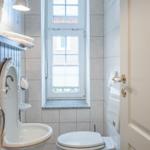 Badezimmer mit Toilette, Waschbecken und Fenster. Holzregal und Spiegel sichtbar.