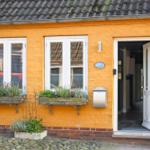 Ein gelbes Haus mit weißen Fenstern und einer offenen Tür. Blumenkästen und ein Briefkasten sind sichtbar.