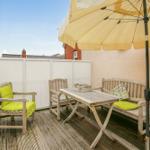 Terrasse mit Holztisch, Bänken und gelbem Sonnenschirm. Holzdielenboden und weiße Umzäunung.