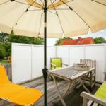 Terrasse mit Sonnenschirm, Holztisch, Stühlen und Liegestuhl. Weiße Umzäunung und Bäume im Hintergrund.