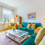 Ein Wohnzimmer mit grünem Sofa, gelber Sessel und Glas-Tisch auf buntem Teppich.