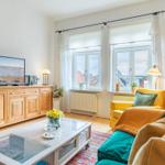 Wohnzimmer mit Fernseher, Couch und Fenstern. Holzmöbel und Blumen sichtbar.