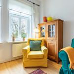 Ein Wohnzimmer mit gelbem Sessel, türkisfarbenem Sofa und Holzschrank. Fenster mit Blick auf Gebäude.
