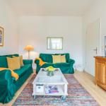 Wohnzimmer mit grünen Sofas, Holzmöbel und Fernseher. Auf dem Teppich steht ein Tisch mit Blumen.