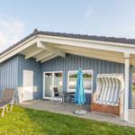 Blauer Ferienhaus mit Terrasse, Liegestühlen und Strandkorb. Grün hinter dem Haus.