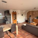 Wohn- und Essbereich mit Küche, Sofa und Holztisch. Terrasse und Kamin sichtbar.