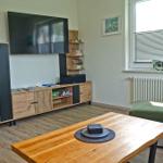 Wohnzimmer mit TV, Holzmöbel und grünem Sofa. Fenster mit Jalousien und Wandbildern.