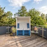 Terrasse mit blauer Sitzbank, Tisch und Pflanzen. Hintergrund: Bäume und Himmel.
