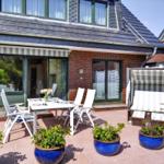 Terrasse mit Tisch, Stühlen und blauen Pflanzkübeln. Hintergrund: Haus mit Glasfront und Dach.