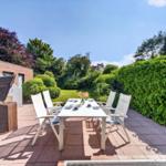 Terrasse mit weißem Tisch und Stühlen, umgeben von grünen Hecken und einem Garten.