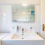 Bathroom with sink, mirror, and shower. White tiles and gold handle.