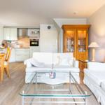 Living and dining area with kitchen. White sofas, glass table, and wooden furniture.