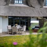 Terrace with table, chairs, and beach chair in front of a thatched-roof house.