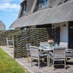 Terrace with table and chairs in front of a house with thatched roof and willow fence.