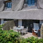 Terrace with table, chairs, and lounge under thatched roof. Surrounded by plants and wooden fence.