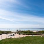 Weitläufiger Strandbereich mit Grünflächen, Bänken und Flaggen unter blauem Himmel.