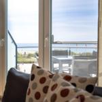 Durch das Fenster ist eine Terrasse mit Tisch und Stühlen sowie Blick aufs Meer zu sehen.