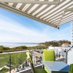 Terrasse mit Sonnenschutz, grünen Stühlen und Blick auf Strand und Wasser.