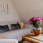 A sofa with cushions is under a sloped ceiling. A table with flowers and a painting are beside it.