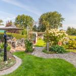 Garden with terrace, flower beds, and wrought iron arch. Cobblestone path leads to the house.
