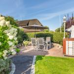 Backyard with seating area, umbrella, and white garden furniture. Lawns and flowers surround the paved area.