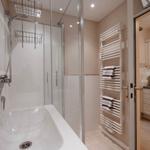 Bathroom with shower, sink, and heater. Kitchen visible in the background.