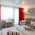 Room with bed, sofa, dining table, and red curtains. Wall art shows lighthouse.