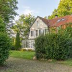 House with red roof, green garden, and gravel path. Surrounded by trees and shrubs.
