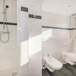 Badezimmer mit Dusche, WC und Bidet. Weiße Fliesen mit dunklen Streifen.