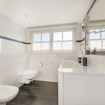 Badezimmer mit Waschbecken, Toilette und Spiegel. Fenster mit Jalousie. Fliesenboden.
