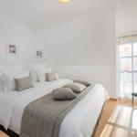 Schlafzimmer mit Doppelbett, weißen Wänden und Holzboden. Fenster mit floralen Vorhängen.