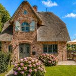 Haus mit strohgedecktem Dach, Gartenbereich mit Sonnenschirm und Liegestühlen, Blumenrabatten und Bäume.