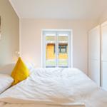 Schlafzimmer mit Bett, gelbem Kissen und Fensterblick auf Holzhaus.