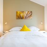Schlafzimmer mit weißem Bett, gelbem Kissen und Wandbild von Blatt