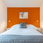 Schlafzimmer mit orangefarbener Wand, Bett mit weißer Bettwäsche und einem Kissen mit Muster.