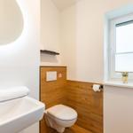 Badezimmer mit Waschbecken, Toilette und Fenster. Holzwand und Spiegel sichtbar.